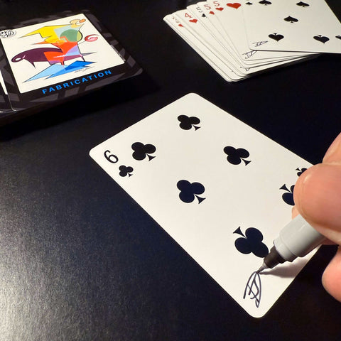 Back of the playing card showing the 6 of clubs with a hand‑signed signature ‘DJ’ in black ink.