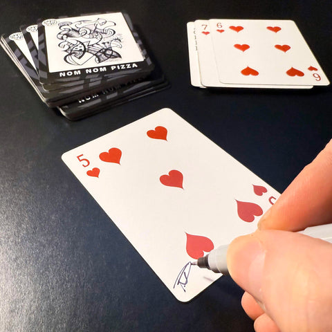 Back of the playing card showing the 5 of hearts with a hand‑signed signature ‘DJ’ in black ink.