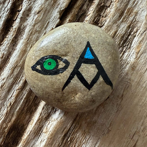 A green eye outlined in black and a stacked AM in the form of a mountain with blue inside the A, resting on a textured wooden surface.