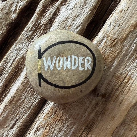 Hand‑painted stone with the word ‘WONDER’ in white within a black oval outline and metallic gold accent, placed on a textured wooden surface.
