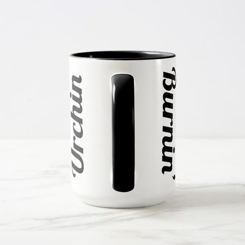 White ceramic coffee mug with a black handle and black interior, featuring a wrap‑around print of the artwork titled “Burnin’ Urchin.“