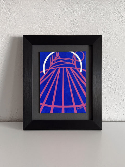 A stylized abstract painting of a tent-like structure with radiating pink lines and a white arc on a vivid blue background; signed 'DARIN' in the lower-right, mounted on a gray mat and framed in black, atop a white shelf, against a white textured wall. 