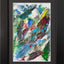 Abstract textured painting with layered brush and palette-knife strokes in blue, green, gray, brown, yellow, and red; matted and framed in black.