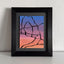 Abstract painting with a black outline of the Delicate Arch against a blue, purple, pink, and orange gradient background; signed, 'DARIN' in lower-right, mounted on a gray mat, framed in black, atop a white shelf and against a textured white wall.