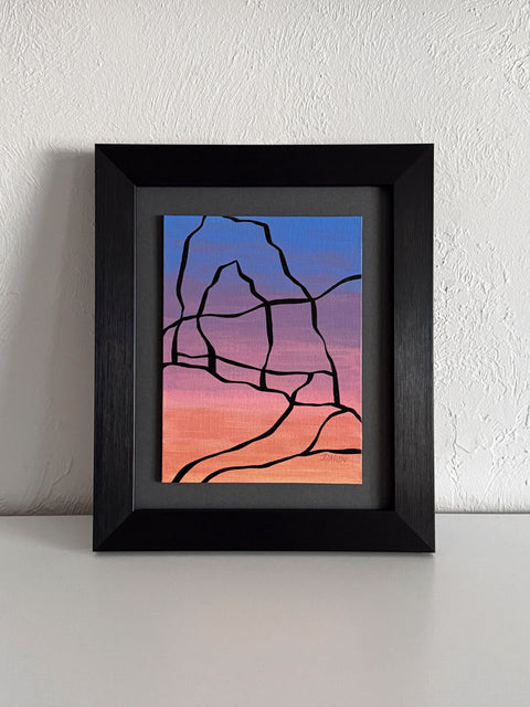 Abstract painting with a black outline of the Delicate Arch against a blue, purple, pink, and orange gradient background; signed, 'DARIN' in lower-right, mounted on a gray mat, framed in black, atop a white shelf and against a textured white wall.